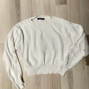 Vit stickad tröja med knappar - Vit stickad tröja från Gina Tricot med rund halsringning och långa ärmar. Tröjan har dekorativa knappar längs ena ärmen och bred ribbstickad mudd vid midjan och ärmsluten. Perfekt för en stilren och trendig look.