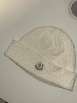 Vit ribbstickad mössa från Moncler - Säljer en stilren vit ribbstickad mössa från Moncler med uppvikt kant och klassisk Moncler-logga framtill. Mössan är gjord i mjukt material och har en clean look som passar soprent till alla outfits. Perfekt för dig som vill ha både värme och hype. Den är använd ett par gånger och är i bra kvalité.