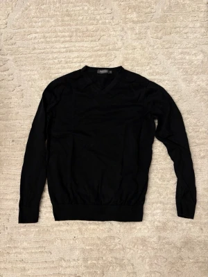 V-ringad tröja Dressmann - Klassisk svart stickad tröja från Dressmann i mjuk bomull. Tröjan har V-ringning, långa ärmar och ribbade muddar vid ärmslut och nederkant. Perfekt att matcha med skjorta eller t-shirt för en clean look.