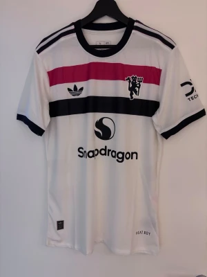Vit Manchester United Adidas tröja L fits M, player edition - Säljer en vit Manchester United matchtröja från Adidas med svarta och rosa detaljer. Tröjan har tryckta loggor, Snapdragon sponsortryck och svarta muddar på ärmarna. Tillverkad i lätt och ventilerande material med HEAT.RDY-teknologi. Är i ny skick, spelar edition/player edition