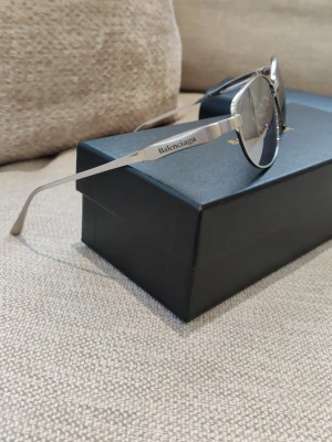 Balenciaga silvriga pilotbågar - Snygga solglasögon från Balenciaga med tunna, silvriga metallbågar.. Glasen är mörkgrå och ger en cool look. Balenciaga-loggan är ingraverad på skalmen för en exklusiv touch. Sitter mycket stabilt.passa de son har medell/stor storlek på glasögon.