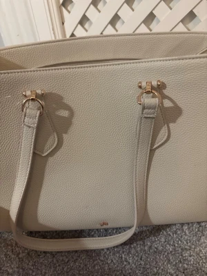 Beige handväska med gulddetaljer - Stilren beige handväska med två långa handtag och snygga guldfärgade metalldetaljer. Väskan har en strukturerad yta i skinnimitation och minimalistisk design. Perfekt för dig som gillar klassiska och eleganta accessoarer.