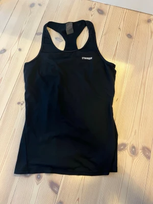 Svart träningstopp från Stronger - Snygg svart träningstopp från Stronger med brottarrygg och diskret logga på bröstet. Tillverkad i stretchigt funktionsmaterial som andas, perfekt för gymmet eller löprundan.