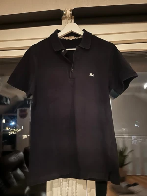 Svart pikétröja från Burberry - Klassisk svart pikétröja från Burberry med en broderad logga på bröstet. Tröjan har krage, knappar framtill och korta ärmar. Perfekt för en stilren och avslappnad look.