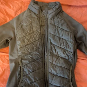 Olivgrön dunjacka från 157 - Snygg olivgrön dunjacka från 157 med quiltade partier och hög krage. Jackan har dragkedja framtill och två fickor på sidorna. Perfekt för kyliga dagar och enkel att matcha med olika outfits.