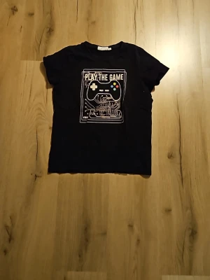 Svart t-shirt med spelkontrolltryck - Cool svart t-shirt med tryck av en spelkontroll och texten 'PLAY THE GAME' på bröstet. T-shirten har korta ärmar och en normal passform. Perfekt för dig som gillar gaming och vill ha en avslappnad stil.