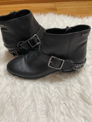 Svarta stövletter från Karl Lagerfeld - Snygga svarta stövletter från Karl Lagerfeld i skinn med rund tå och dekorativa silverfärgade spännen och kedjedetaljer vid hälen. Dragkedja på insidan för enkel på- och avtagning. Perfekta för att lyfta din outfit.