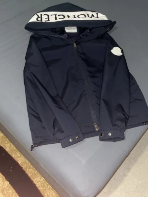 Mörkblå Moncler vindjacka med huva - Snygg mörkblå vindjacka från Moncler med stor logga på ärmen och huva med vit insida och Moncler-tryck. Jackan har dragkedja framtill och tryckknappar vid ärmsluten. Perfekt för dig som vill ha en stilren och exklusiv look.