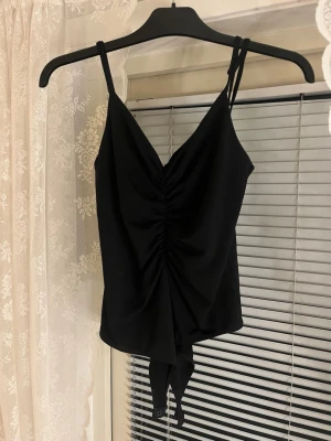 Svart body med srunch - Snygg svart body med smala axelband och rynkdetalj framtill. Djup V-ringning och knäppning i grenen. Perfekt att styla till jeans eller kjol för en trendig look. Materialet känns mjukt och stretchigt för skön passform.