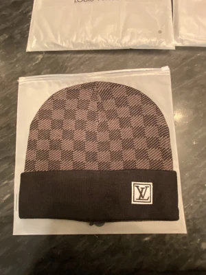Louis Vuitton svart rutig mössa - Snygg svart mössa från Louis Vuitton med klassiskt rutigt mönster och vikt kant. LV-logga framtill på kanten ger en exklusiv touch. Perfekt accessoar för att lyfta din stil under kyliga dagar.