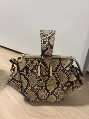 Beige handväska med ormmönster - Snygg handväska i beige och svart med ormmönster och guldfärgade detaljer. Väskan har en unik geometrisk form och dragkedja upptill. Perfekt accessoar för att lyfta din outfit.