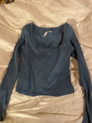 Blå ribbad topp från H&M Divided - Snygg blå långärmad topp från H&M Divided med ribbad struktur och rundad urringning. Perfekt att styla med jeans eller kjol för en trendig look. Materialet är mjukt och stretchigt för extra komfort.
