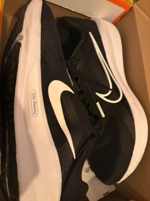 Nike Downshifter 13 svart/vit sneakers - Nike Downshifter 13 sneakers i svart och vitt med stor vit swoosh på sidan och mesh-överdel. Skorna har snörning, platt sula och rund tå. Yttersulan är svart med mönstrad struktur och vit swoosh. Perfekta för löpning och träning, med lätt och ventilerande syntetmaterial. Testade ute en gång men är för stora!  Storlek 43 Skicket är nytt & syns på bilderna, lite smutsiga under pga testade!  Köpta för 699kr  Skickas i originalförpackning självklart!