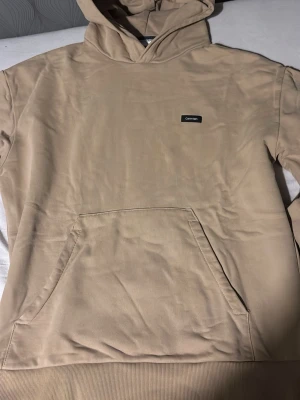 Beige hoodie från Calvin Klein - Snygg beige hoodie från Calvin Klein med liten logotyp på bröstet. Hoodien har känguruficka framtill och en klassisk huva. Tillverkad i mjuk bomull för skön komfort och har en relaxed passform.