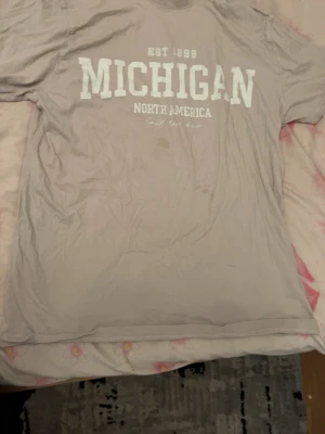 Beige Michigan t-shirt med tryck - Snygg beige t-shirt med stort vitt 'MICHIGAN' tryck på bröstet och texten 'North America'. Klassisk passform och korta ärmar, perfekt för en avslappnad stil. Tillverkad i mjuk bomull som känns skön mot huden.
