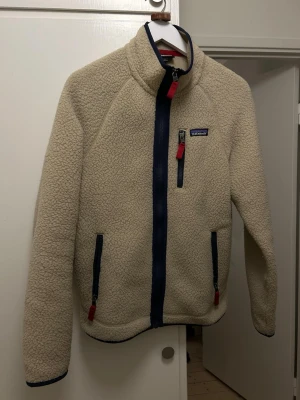 Beige teddyjacka från Patagonia - Mysig beige teddyjacka från Patagonia med marinblå detaljer och dragkedjor. Jackan har hög krage, tre fickor med dragkedja och röd zip-puller. Perfekt för kalla dagar och ger en avslappnad, trendig look.