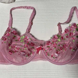 Rosa spets-bh med blommor från Lounge - Söt rosa bh från Lounge med transparent mesh och broderade blommor i rosa, rött och grönt. Bh:n har bygel, justerbara axelband och en liten rosett framtill. Perfekt för dig som gillar romantiska detaljer och blommigt mönster. Aldrig använd, endast provad. Säljs pga för stor storlek. 300kr