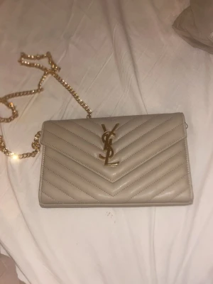 Beige YSL kuvertväska i skinn - Snygg beige kuvertväska från YSL i skinn med quiltad design och guldigt YSL-emblem framtill. Väskan har en elegant kedjerem i guld som gör den enkel att bära över axeln. Perfekt accessoar för att lyfta din outfit.