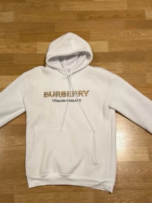 Burberry hoodie - Snygg vit hoodie från Burberry med klassisk logga i beige och svart på bröstet samt texten 'London England' under. Hoodien har huva med dragsko och en stor magficka framtill. Tillverkad i mjukt material som känns skönt mot huden. Perfekt för en clean och stilren look.