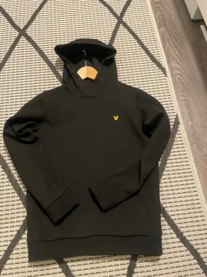 Svart hoodie från Lyle & Scott - Snygg svart hoodie från Lyle & Scott med klassisk gul logga på bröstet. Hoodien har huva och en stor magficka framtill. Tillverkad i mjukt bomullsmaterial som är skönt att bära. Perfekt för en avslappnad och stilren look.