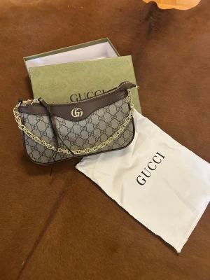 Gucci GG Supreme handväska med kedja - Snygg handväska från Gucci i beige och brunt GG Supreme-mönster med guldfärgad kedjedetalj och GG-logo framtill. Väskan har dragkedja upptill och detaljer i brunt skinn. Perfekt accessoar för att lyfta din outfit.
