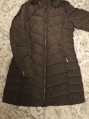  lång dunjacka med dragkedja - Snygg brun lång dunjacka med quiltad design och dragkedja framtill. Jackan har två snedställda fickor med dragkedja och en hög krage med fuskpälsdetalj. Perfekt för kalla dagar och ger en stilren look.