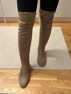 Beige overknee stövlar med snörning - Snygga beige overknee stövlar i mocka-look med rund tå och snörning baktill upptill. Stövlarna har platt sula och sitter tight längs benet för en stilren siluett. Perfekta att styla med kjol eller klänning.