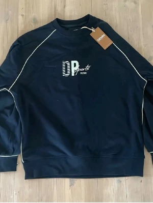 Mörkblå sweatshirt från DP Sports - Mörkblå sweatshirt från DP Sports med vit piping längs ärmarna och axlarna. Tryckt logga på bröstet och rund halsringning. Tröjan har en avslappnad passform och ribbade muddar vid ärmslut och nederkant. Perfekt för en sportig och casual stil.