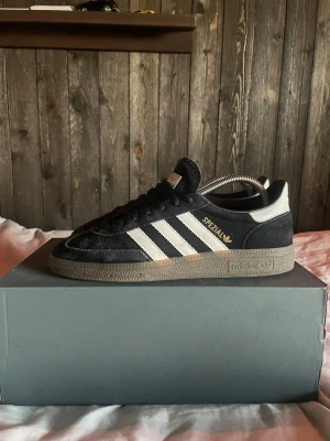 Adidas Spezial svarta sneakers mocka - Säljer ett par Adidas Spezial sneakers i svart mocka med vita detaljer och klassiska tre ränder på sidan. Skorna har snörning, gummisula och guldfärgad Spezial-text på sidan. Perfekta för dig som gillar retrostil och streetwear.