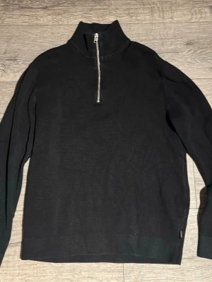 Svart stickad tröja med halv dragkedja - Svart stickad tröja med hög krage och halv dragkedja framtill. Tröjan har lång ärm och en stilren, minimalistisk design. Perfekt för lager på lager och passar till det mesta. Materialet ser ut att vara mjukt och bekvämt.