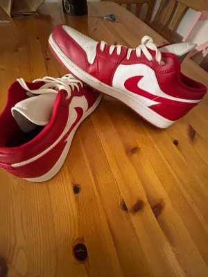 Nike Air Jordan 1 Low röd/vit US13 köpt för 2700 kr - Säljer ett par Nike Air Jordan 1 Low i rött och vitt skinn. Klassisk siluett med vit bas, röda overlays och ikonisk Swoosh på sidan. Skorna har snörning och perforerad tå för extra ventilation. Perfekta för dig som vill sticka ut med en tidlös sneaker.