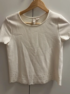Vit topp med broderad kant H&M - Söt vit topp från H&M med korta ärmar och rund halsringning. Nederkanten har broderade detaljer och vågig avslutning som ger en romantisk touch. Tillverkad i ett strukturerat tyg som känns mjukt mot huden.