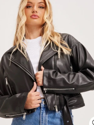 Svart croppad skinnjacka med bälte - Snygg svart skinnjacka i bikerstil med croppad passform, breda slag och flera silvriga dragkedjor. Jackan har bälte i midjan och detaljerade spännen vid ärmslut. Perfekt för en edgy look.