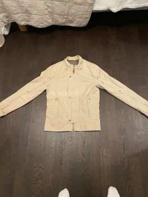 Beige vindjacka med ståkrage - Snygg beige vindjacka med ståkrage och tryckknappar framtill. Jackan har långa ärmar, två fickor med knappstängning och dragkedja. Perfekt för dig som vill ha en stilren och enkel jacka till vardags.