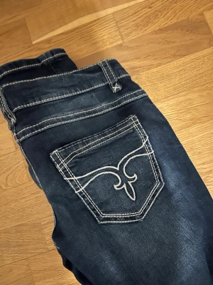 Mörkblå bootcut jeans med kontrastsömmar - Snygga mörkblå jeans med vita kontrastsömmar och unik broderad bakficka. Bootcut-modell som ger en schysst siluett och passar perfekt till sneakers eller boots. Klassisk femficksdesign.
