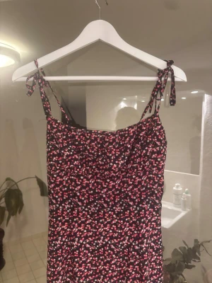 Blommig kort klänning med knytband - Söt kort klänning med smala axelband som knyts upptill. Klänningen har ett blommigt mönster i rosa, vitt och rött på svart botten. Lätt utsvängd nederdel och figurnära passform. Perfekt för varma dagar.