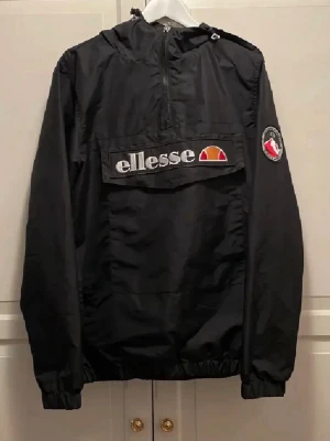 Svart vindjacka från Ellesse - Snygg svart vindjacka från Ellesse med stor logga på bröstet och patch på ärmen. Jackan har huva, half zip och en stor ficka framtill. Perfekt för dig som vill ha en sportig och stilren look.