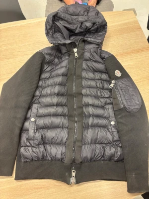 Svart dunjacka med huva från Moncler - Snygg svart dunjacka från Moncler med quiltad front, stickade ärmar och huva. Jackan har dragkedja framtill, fickor med tryckknappar och en extra ficka med dragkedja på ärmen. Perfekt för kalla dagar och riktigt stilren look. Priset kan diskuteras!😁