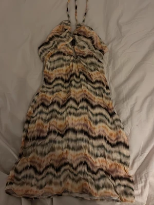 Mönstrad halterneckklänning i pastell - Säljer en kort halterneckklänning med randigt mönster i beige, svart och orange toner. Klänningen har dekorativa träpärlor framtill och tunna knytband i nacken. Materialet känns lätt och luftigt, perfekt för varma dagar.