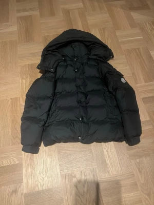 Svart pufferjacka från Moncler - Säljer en svart pufferjacka från Moncler med huva och Moncler-logga på ärmen. Jackan har quiltad design, långa ärmar och dragkedja framtill. Perfekt för kalla dagar och riktigt snygg till streetwear-stilen.