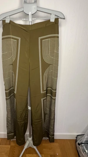 Olivgröna satinbyxor från H&M M - Snygga olivgröna satinbyxor från H&M i storlek M. Byxorna har ett unikt vitt grafiskt mönster längs benen och en rak, loose passform. Perfekta för dig som vill sticka ut med coola detaljer och bekvämt material.