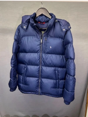 Mörkblå pufferjacka från Polo Ralph Lauren - Snygg mörkblå pufferjacka från Polo Ralph Lauren med huva och dragkedja framtill. Jackan har det klassiska lilla logobroderiet på bröstet och en quiltad design som ger en riktigt fet look. Perfekt för kalla dagar när du vill hålla stilen.