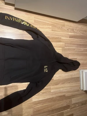 Svart hoodie från Emporio Armani - Snygg svart hoodie från Emporio Armani med guldfärgat EA7-tryck på bröstet och stort Emporio Armani-tryck längs ena ärmen. Klassisk känguruficka framtill och justerbar huva med snören. Perfekt för dig som gillar exklusiv streetwear.