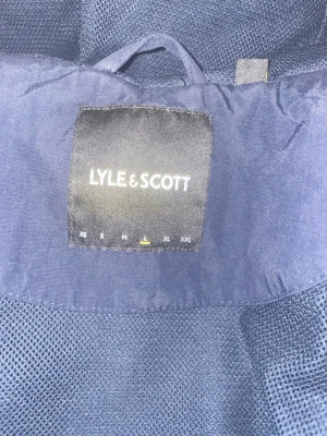 Marinblå jacka från Lyle & Scott - Snygg marinblå jacka från Lyle & Scott med meshfoder och klassisk design. Perfekt för dig som vill ha en stilren och enkel jacka med diskreta detaljer. Passar till många olika outfits och har en sportig känsla. Nackdel: vänster ficka saknar knapp. 