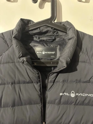 Svart dunväst från Sail Racing - Snygg svart dunväst från Sail Racing med diskret logga på bröstet och ryggen. Köpte den för 1 år sedan för 2200 och nu har jag växt ur den storlek medium och passar nån 175-185 typ.