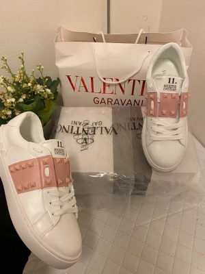 Valentino Rockstud Untitled sneakers - Snygga vita sneakers från Valentino med rosa band och ikoniska nitar över vristen. Modellen heter Rockstud Untitled och har en chunky sula, snörning och detaljer i skinn. Perfekta för dig som vill sticka ut med exklusiv design och coola detaljer.