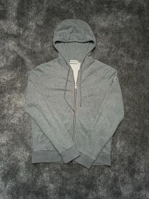 Moncler hoodie - Snygg grå hoodie från Moncler med dragkedja och klassisk huva. Tröjan har ribbade muddar och Moncler-logga på ärmen. Dragkedjan har en diskret röd, vit och blå detalj. Perfekt för en avslappnad och stilren look.