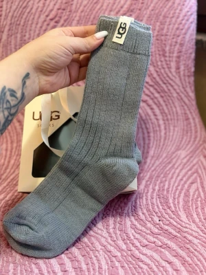 Grå ribbade strumpor från UGG - Säljer ett par grå ribbade strumpor från UGG. De är mjuka och sköna, perfekta för att hålla fötterna varma. Klassisk UGG-logga upptill och snygg ribbad struktur. Passar dig som gillar stilrena och bekväma strumpor.