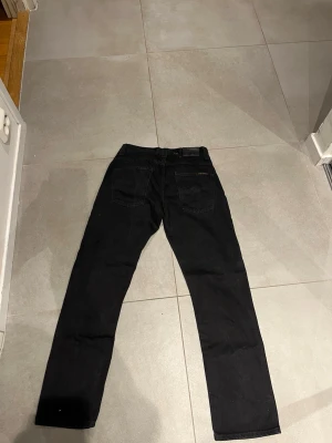 Svarta jeans från Nudie Jeans - Säljer ett par svarta jeans från Nudie Jeans med klassisk femficksmodell och raka ben. Jeansen har svarta sömmar och Nudie Jeans-logga på bakfickan. Perfekta för en clean och stilren look.