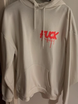 Vit hoodie med röd graffiti-text - Säljer en vit hoodie med huva och känguruficka. På bröstet finns en lite mindre röd graffiti-inspirerad text med ordet 'FUCK' och en stor text på baksidan. Perfekt för en streetwear-look. Hoodien är väldigt nopprig, och har en fläck vid vänster framsida axel.